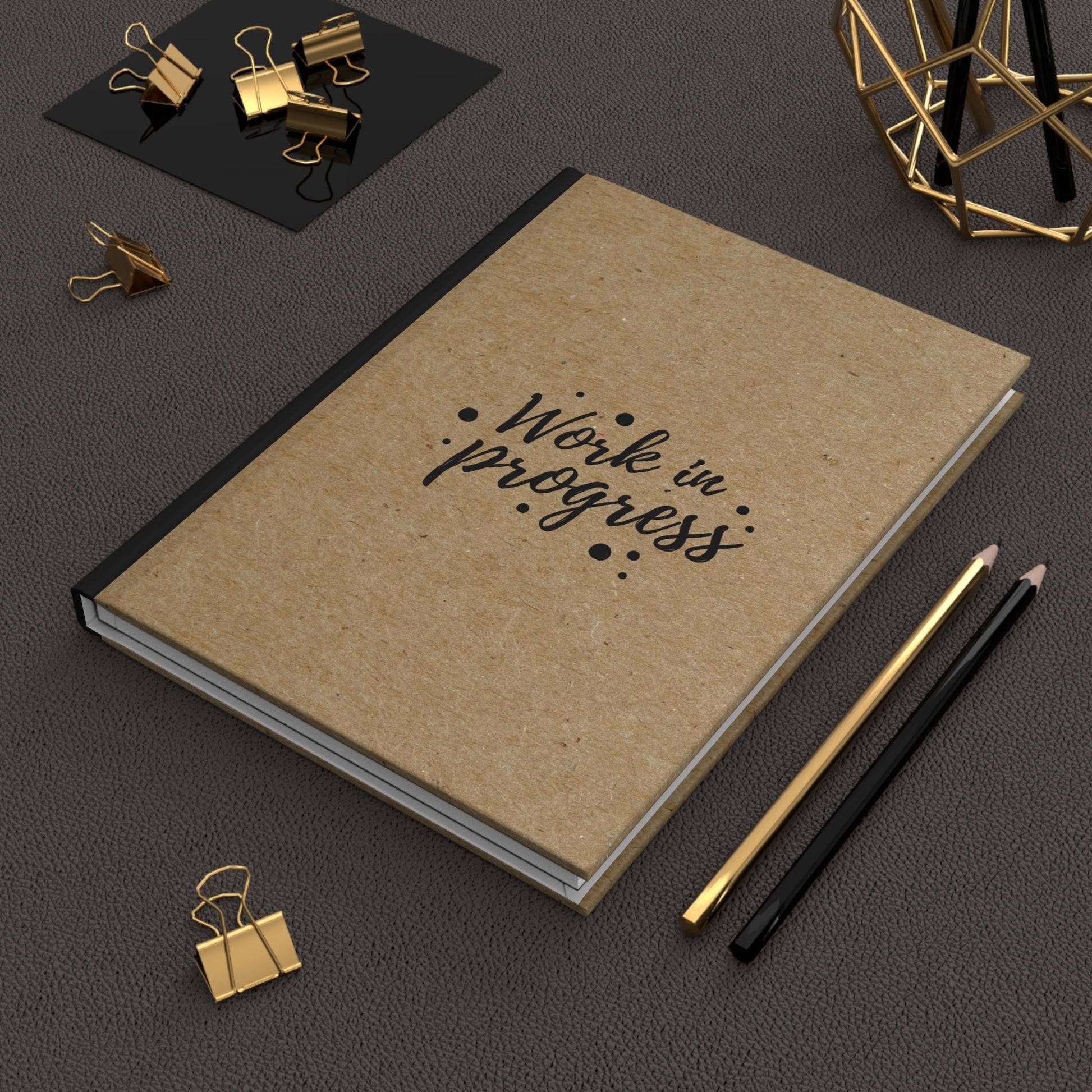 Work in Progress Hardcover Journal with kraft paper cover, black spine, and decorative text, placed on dark surface with pencils and gold binder clips nearby