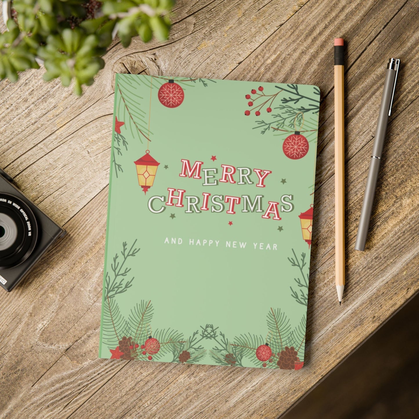 Softcover journal with Merry Christmas and Happy New Year design on green satin finish cover placed on wooden table with pencil and pen