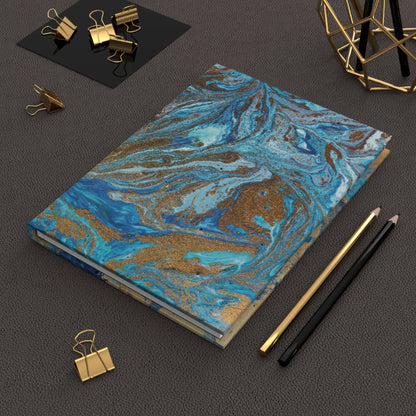 Hardcover notebook with royal blue and gold swirl design on textured surface with pencils and gold clips nearby