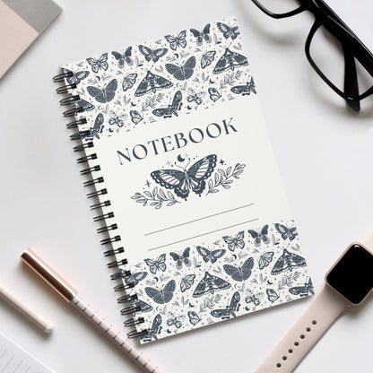 Butterfly spiral notebook with nature-inspired black and white butterfly cover and durable spiral binding on a white desk with glasses, smartwatch, and pens