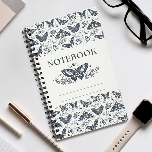 Butterfly spiral notebook with nature-inspired black and white butterfly cover and durable spiral binding on a white desk with glasses, smartwatch, and pens